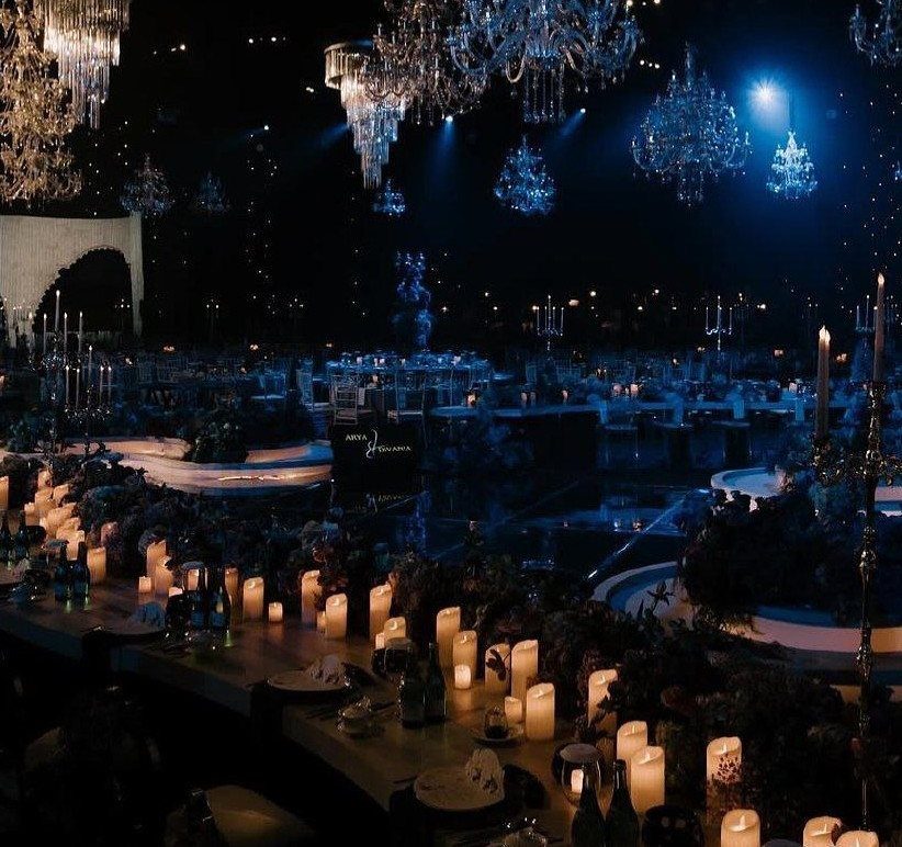 Lavish event tables with glowing candles under crystal chandeliers and deep blue lighting.