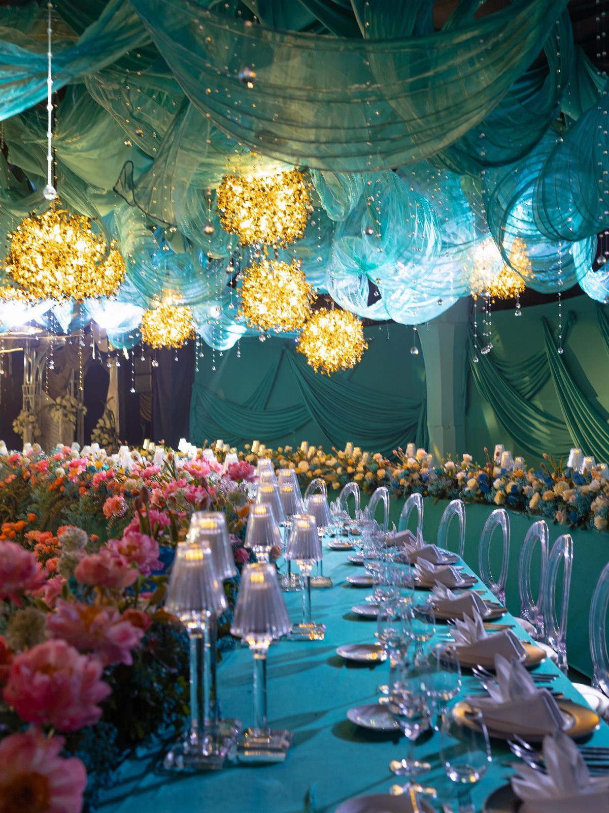 Teal blue banquet table with coral flowers and glowing gold spherical chandeliers.