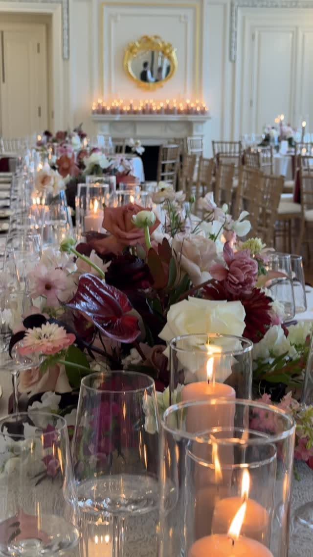 Burgundy and blush floral centerpieces with glowing candles on a long wedding table.