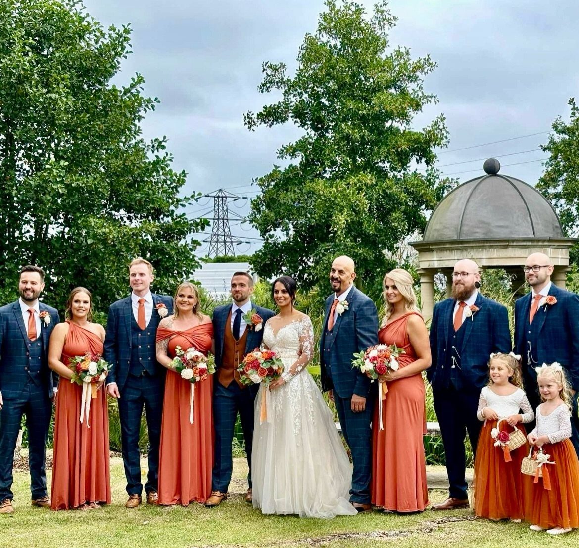 Wedding party in navy suits and burnt orange dresses holding orange floral bouquets.
