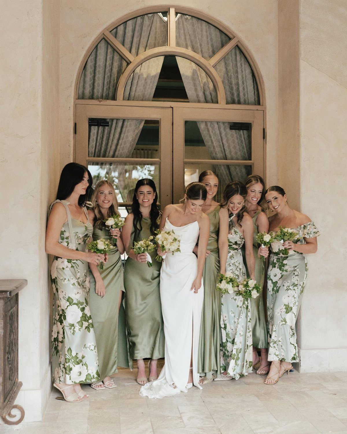Bride in white gown with bridesmaids wearing sage green dresses and floral bouquets.