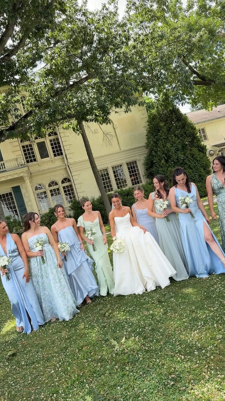 Bride in white gown with bridesmaids wearing blue and green dresses outdoors.