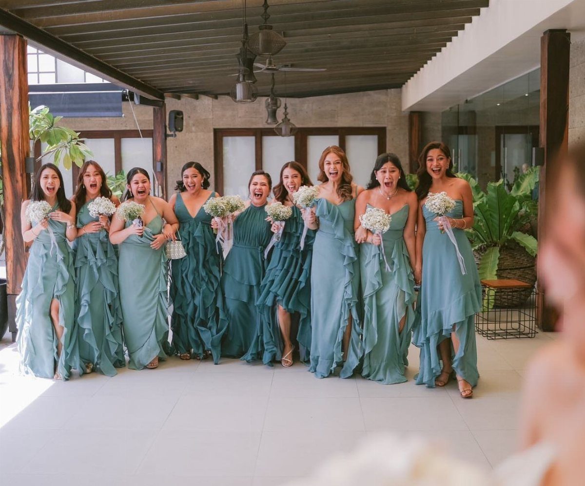 Bridesmaids in teal and sage green gowns holding white floral bouquets outdoors.