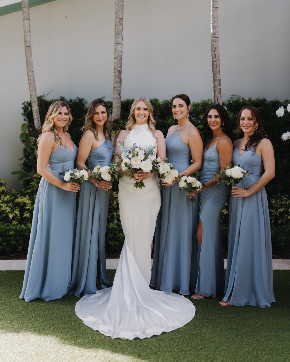 Bride in white gown surrounded by bridesmaids wearing dusty blue dresses with bouquets.