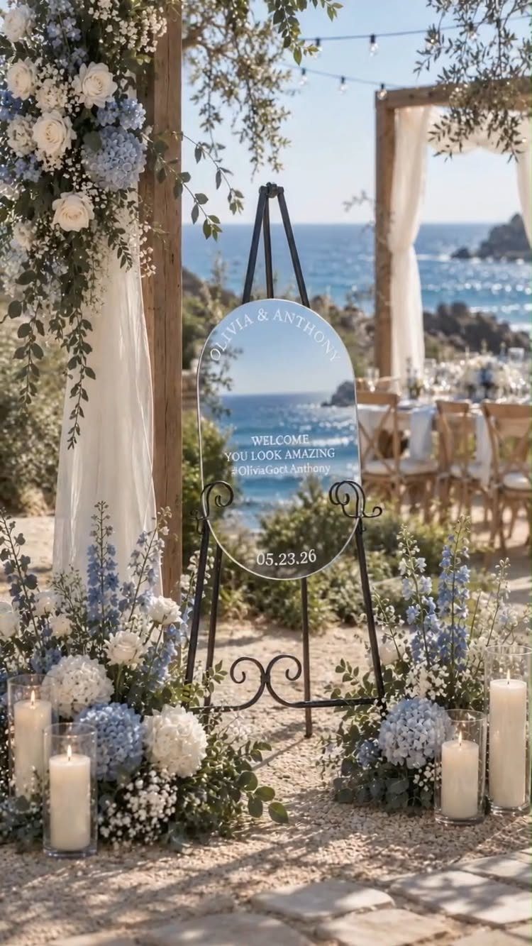 Mirrored wedding welcome sign surrounded by blue and white flowers on a sandy beach.