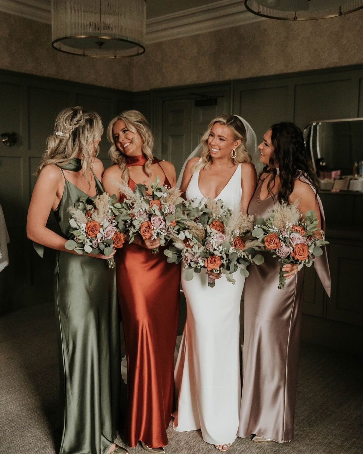 Bride and bridesmaids in olive, rust, and mauve dresses holding matching floral bouquets.