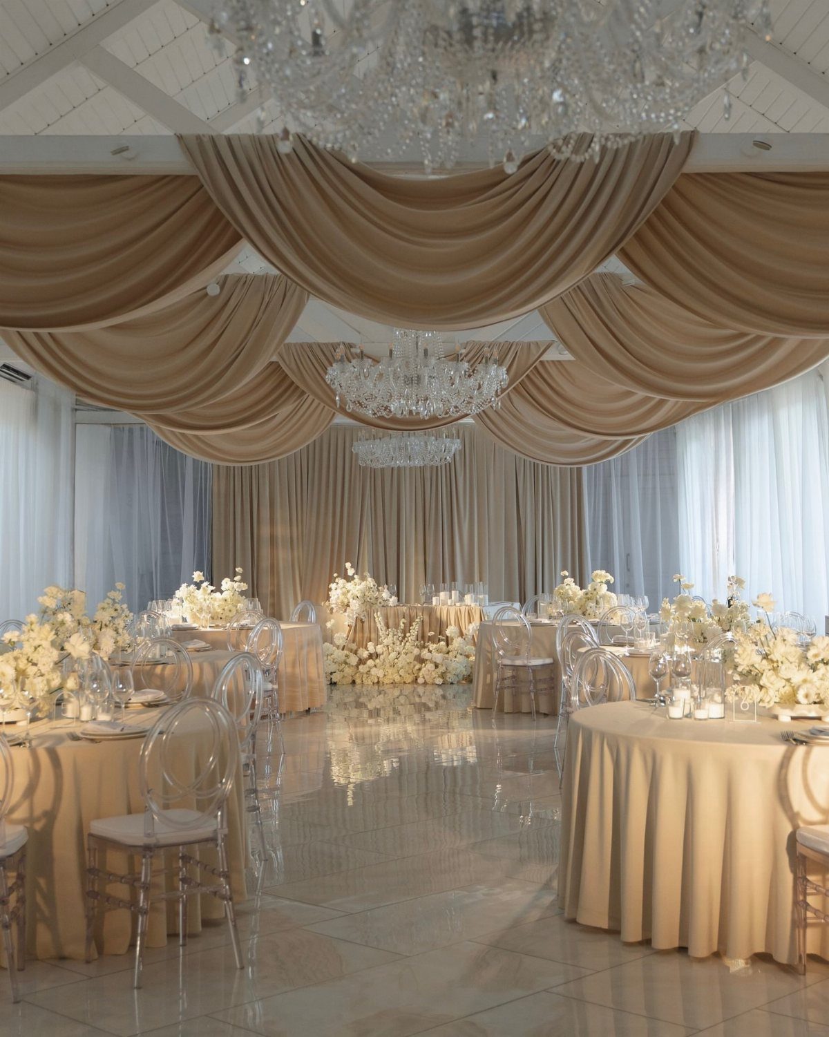 Champagne ballroom with draped ceilings, matching linens, and beige floral centerpieces.