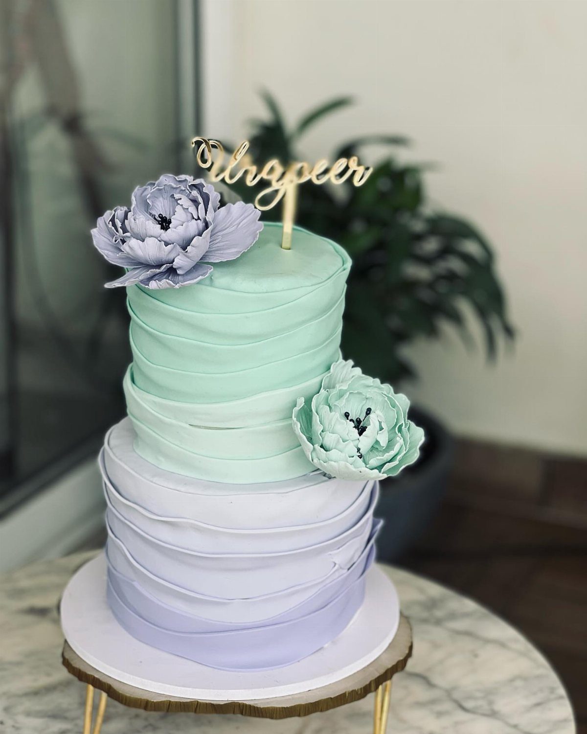 Two-tier ruffled cake in mint green and lavender with floral decorations and gold topper.