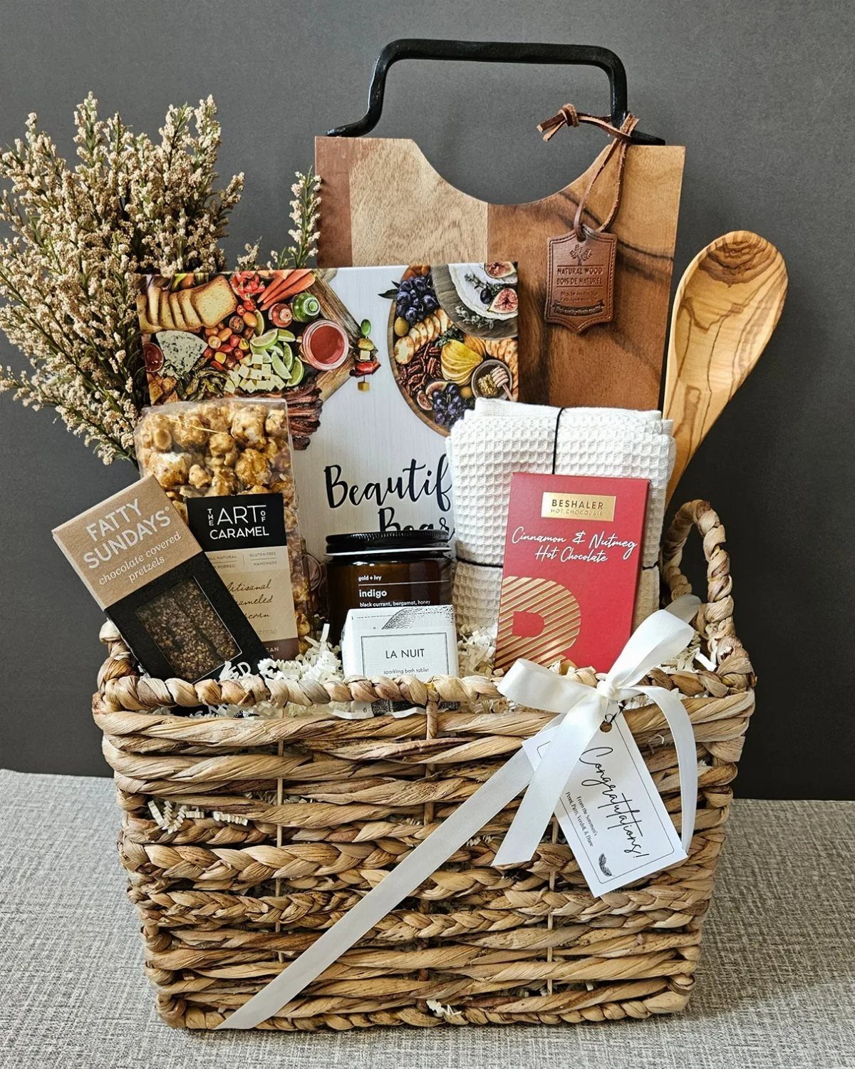 Woven wicker gift basket filled with gourmet treats, a cutting board, and a cookbook.