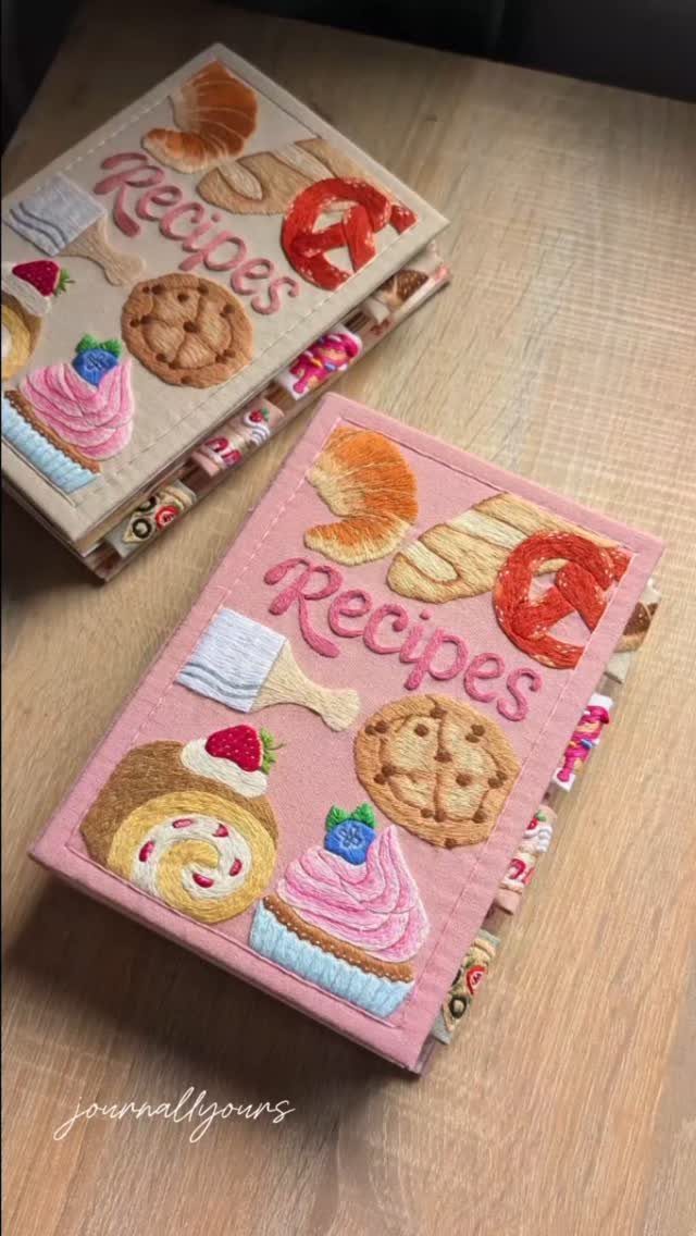 Two fabric recipe books with embroidered food illustrations on a wooden table.