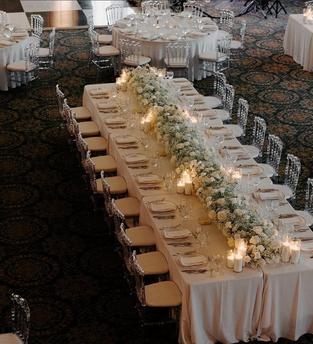 Long wedding head table with white floral runner, candles, and clear ghost chairs.