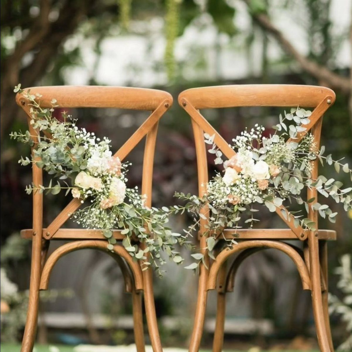 Two wooden cross-back chairs decorated with white roses and eucalyptus greenery for a wedding.