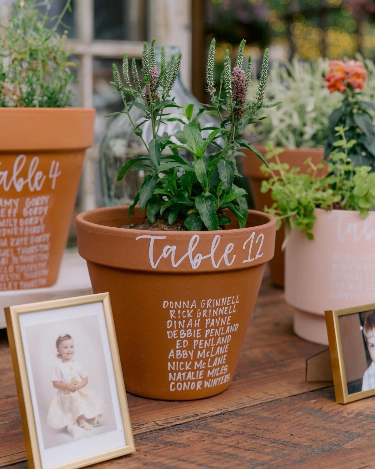 Terracotta pots with table numbers and guest lists next to gold framed photos.