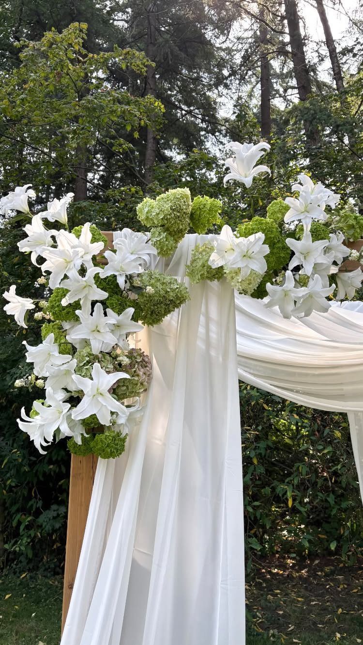 Wooden wedding arch with white fabric draping and white lily floral arrangements outdoors