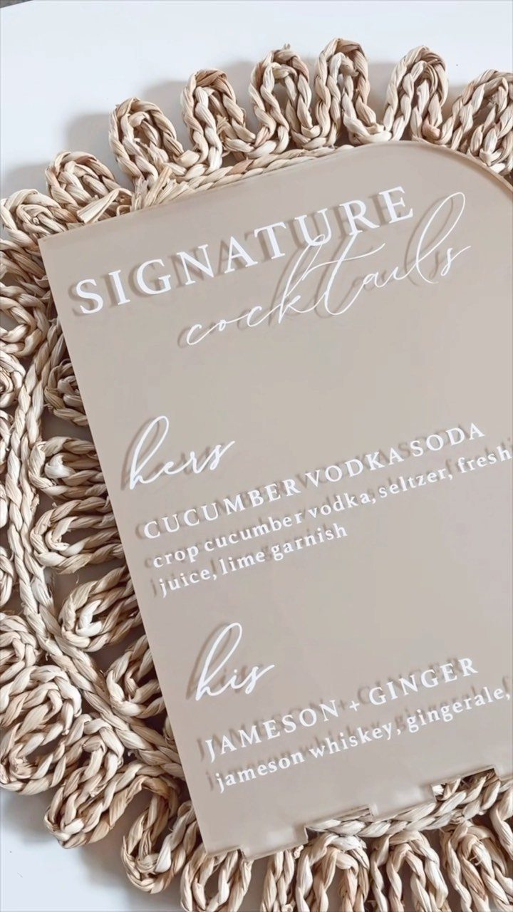 Acrylic signature cocktail sign with white text resting on a woven straw mat.