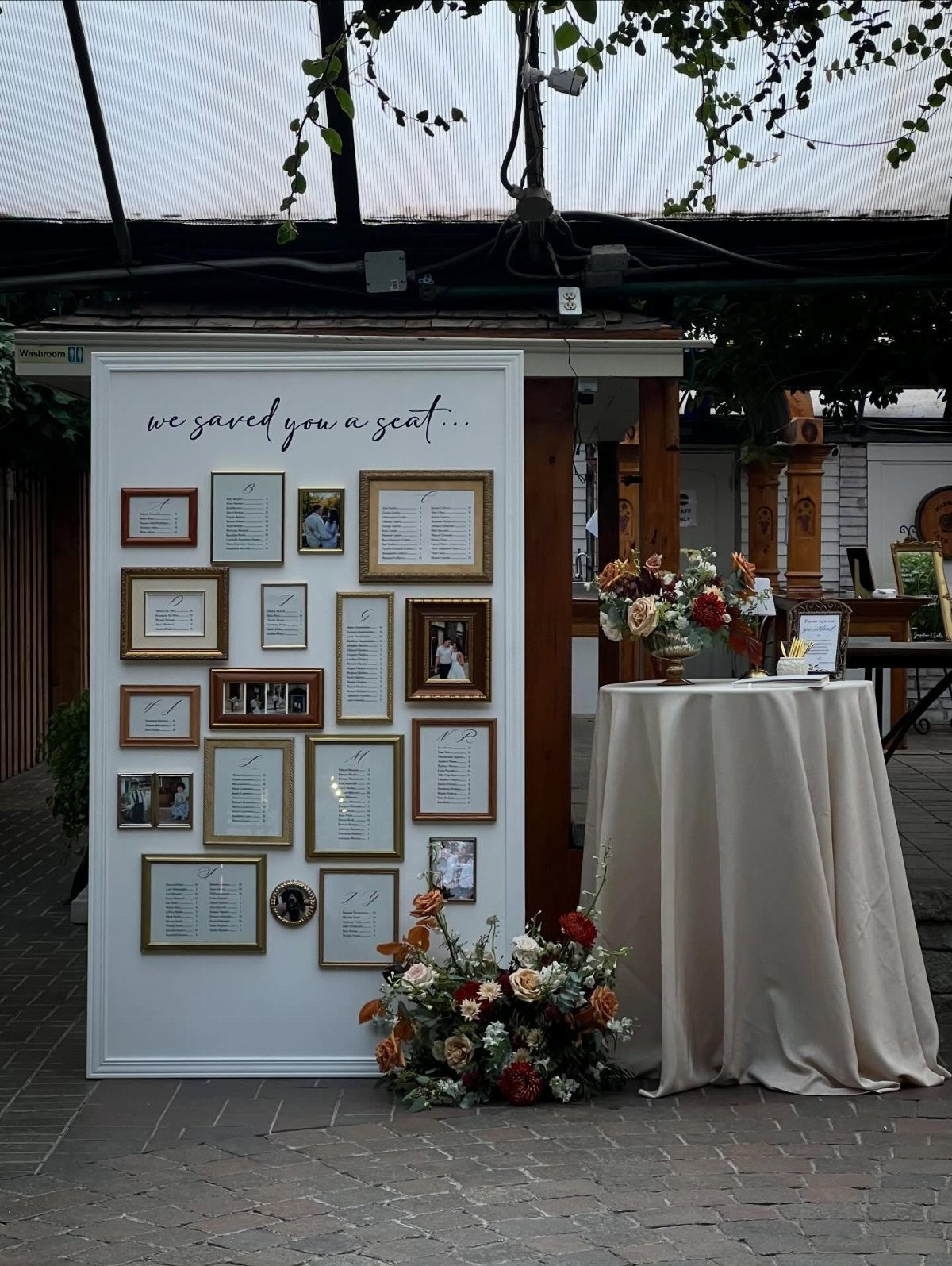 White display board with gold-framed seating arrangements and photos next to a floral table.