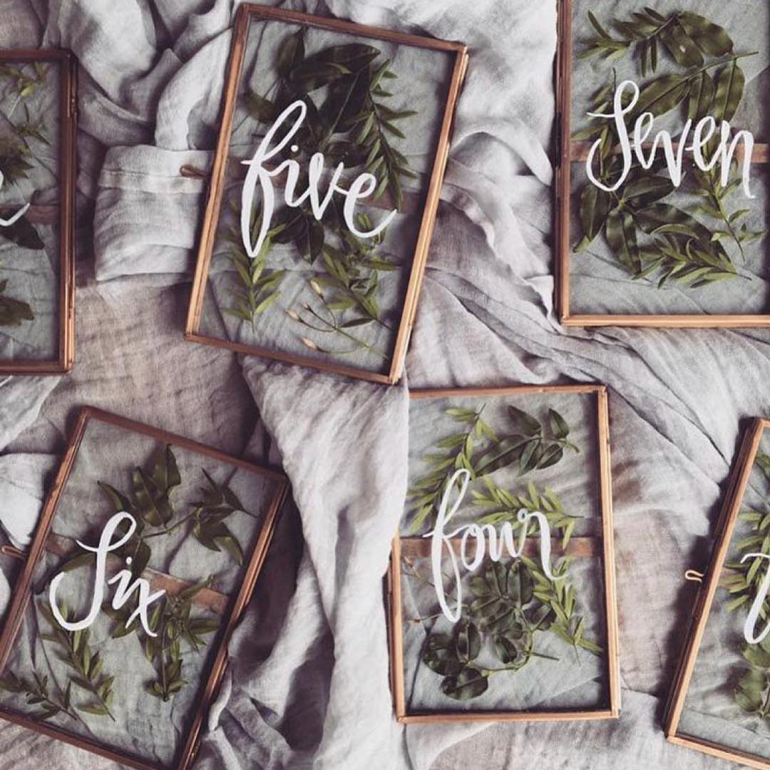Glass table number frames with green leaves and white cursive text on grey fabric