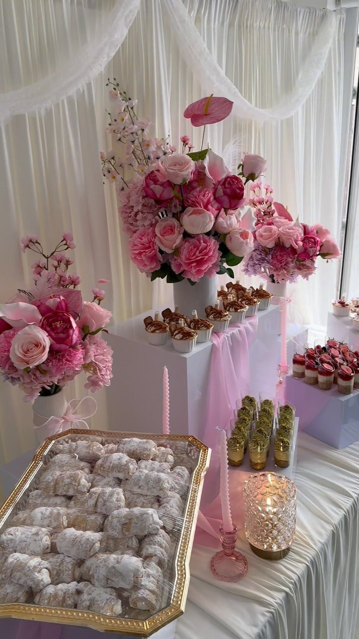 Dessert table with powdered pastries, cupcakes, pink flowers, and white draped fabric backdrop.