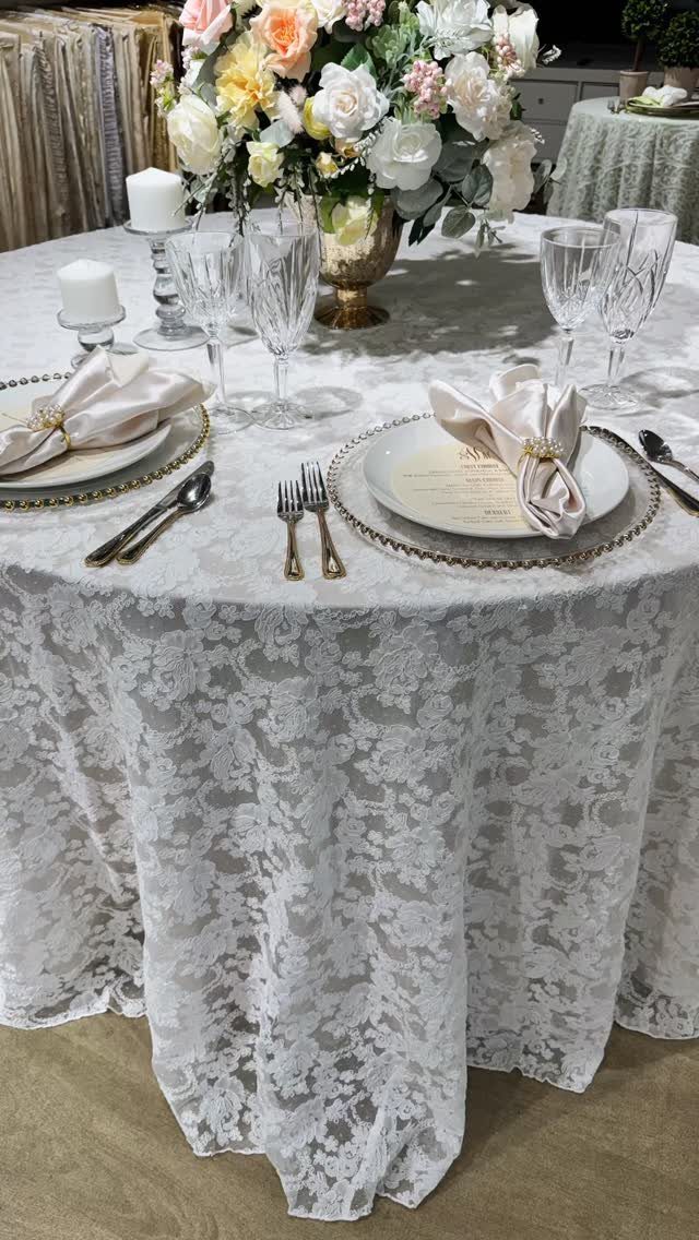 Round wedding table with white lace tablecloth, gold plates, and floral centerpiece.