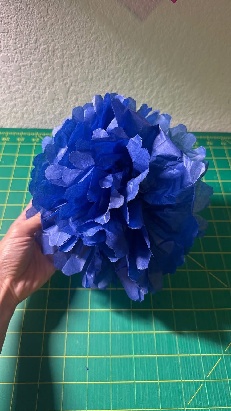 Blue tissue paper pom-pom flower held by a hand over a green mat.