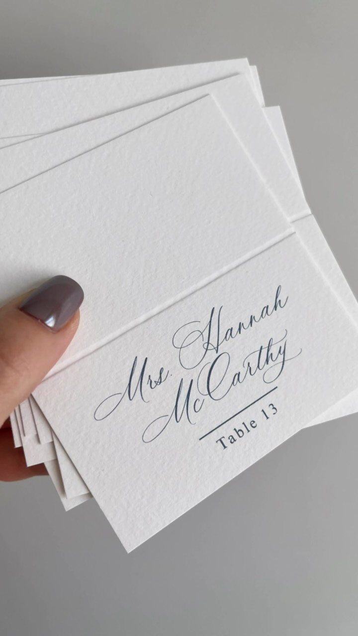 White textured name cards with navy blue calligraphy and table number held by hand.
