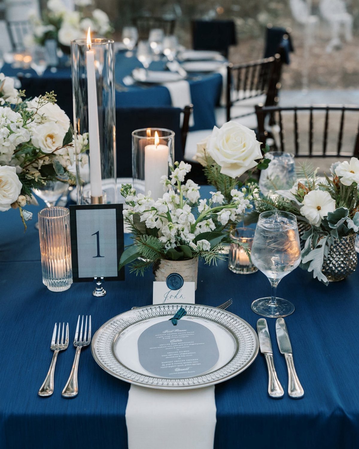 Navy blue wedding table setting with white roses, candles, and silver dinnerware.