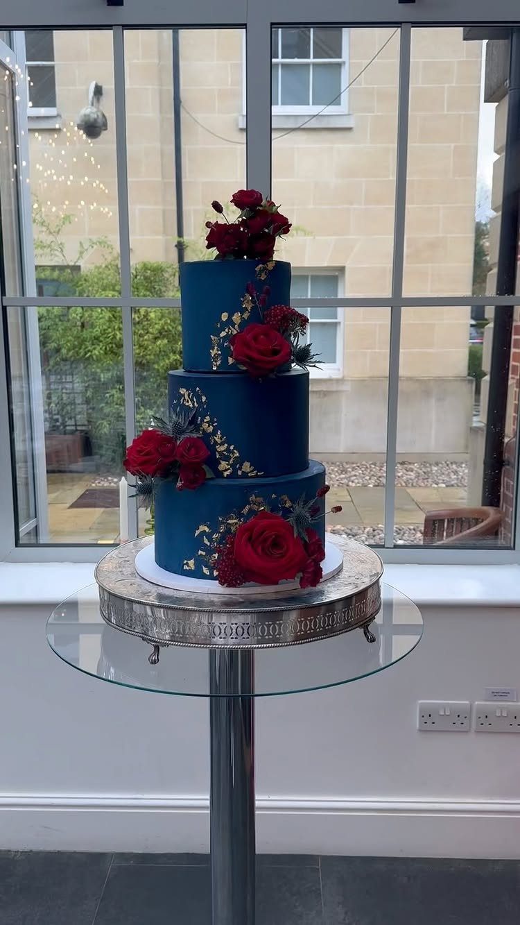 Three-tiered navy blue wedding cake with red roses and gold leaf accents.