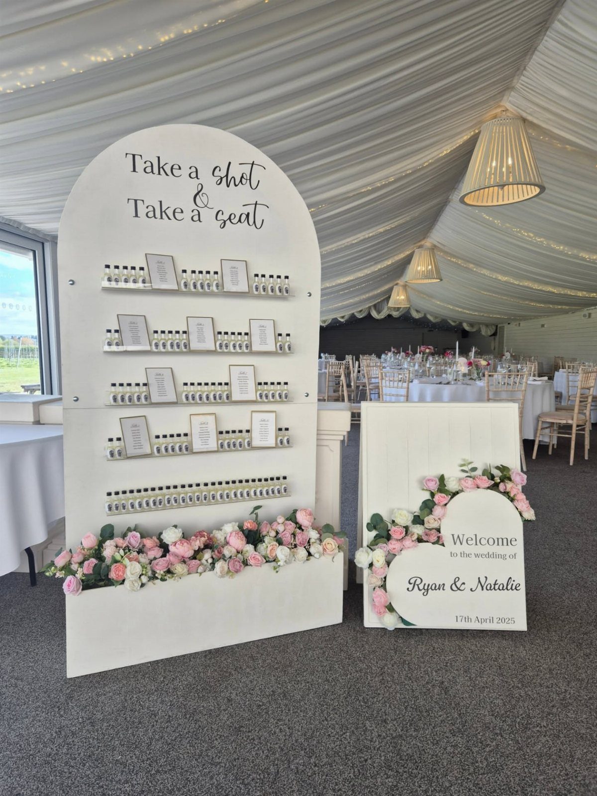 White arched favor table with small bottles and pink floral arrangements in a tent