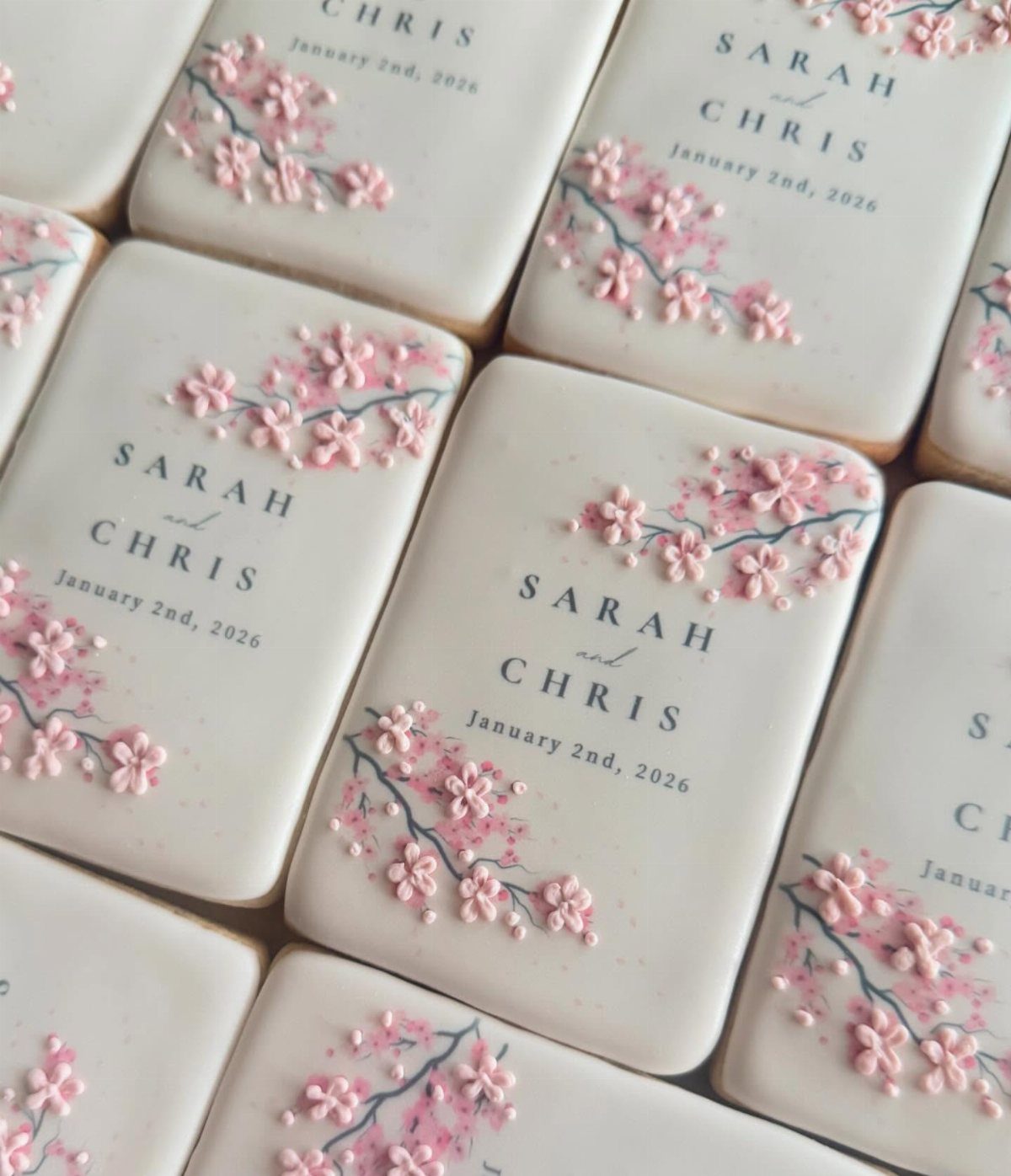 Rectangular sugar cookies with white icing, pink cherry blossoms, and wedding names.