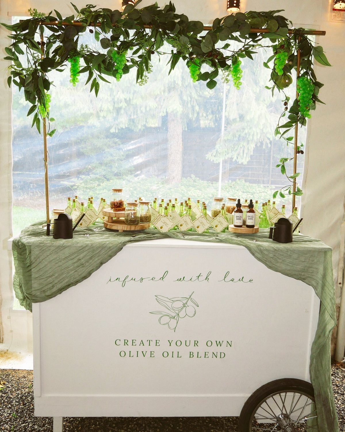 Olive oil blending station on a white cart with greenery and glass bottles.