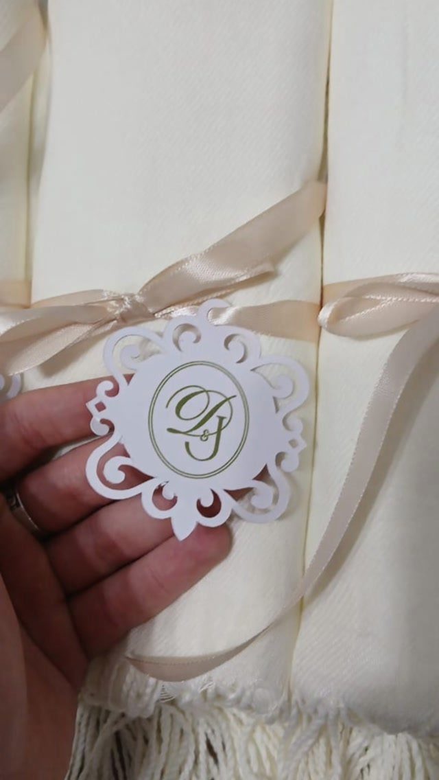 Cream-colored pashmina shawls rolled and tied with ribbons and personalized monogram tags.