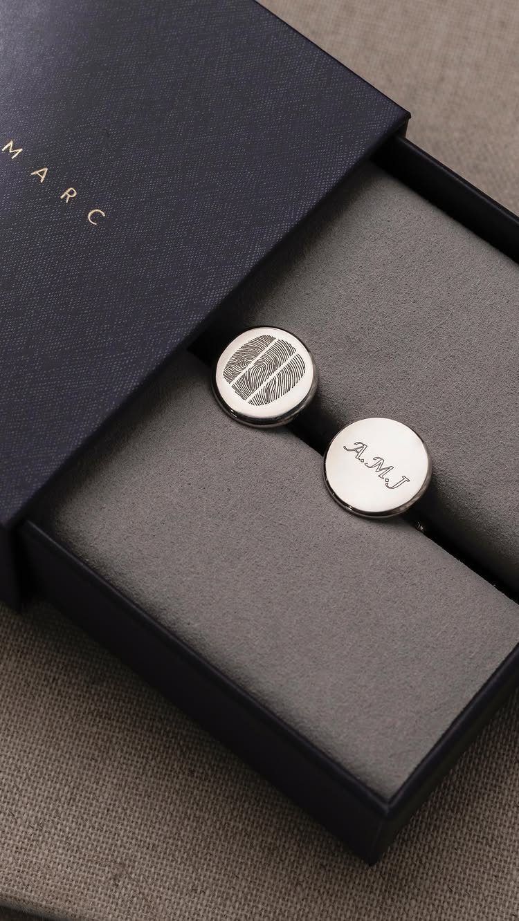 Silver circular cufflinks with fingerprint and initial engravings in a blue jewelry box.