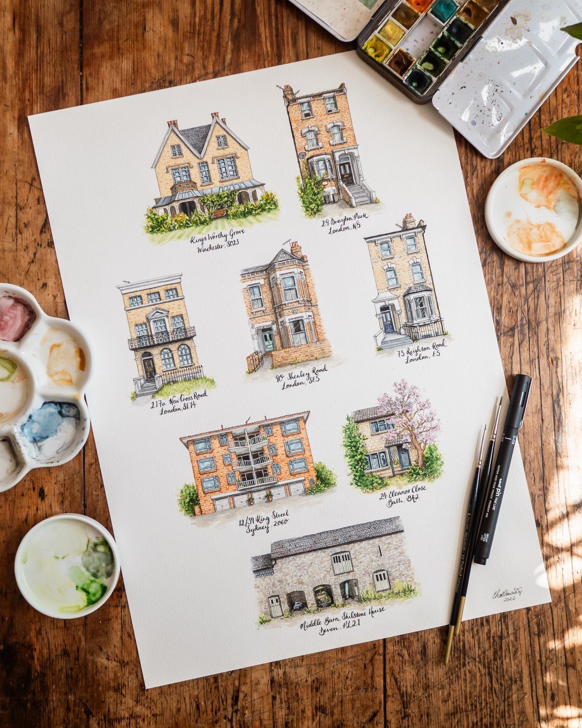 Watercolor paintings of various houses on a white page surrounded by art supplies.