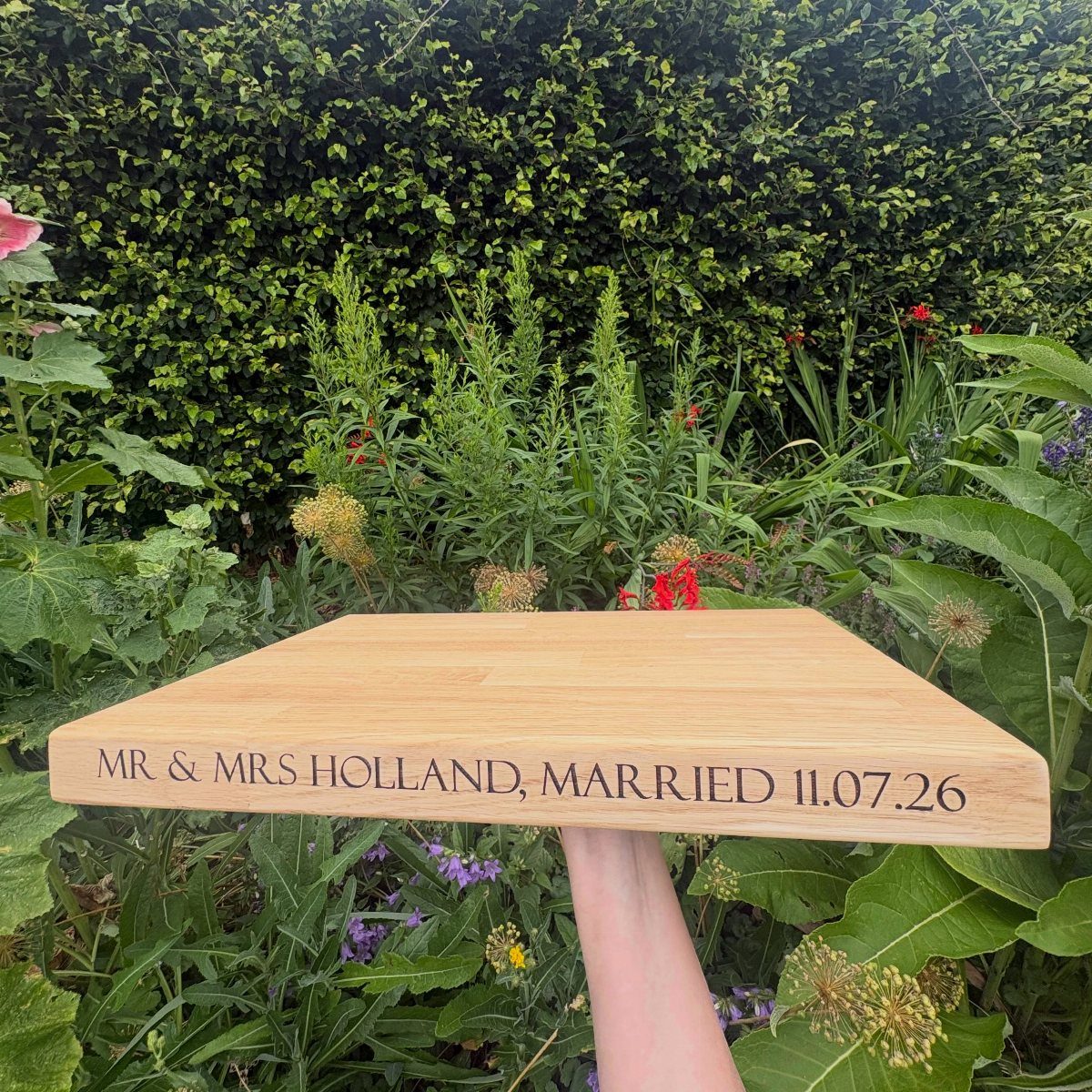 Engraved wooden cutting board held against a green garden background with wedding text.