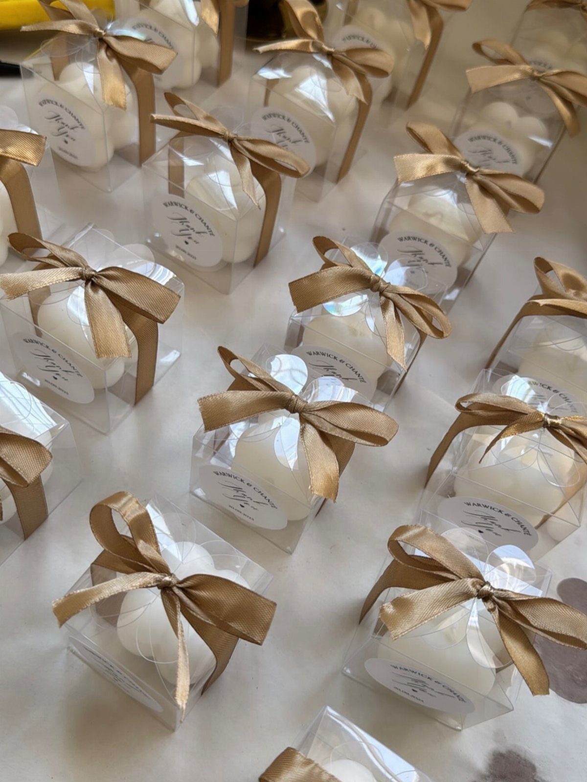 Small white scented candles in clear boxes with gold ribbons and personalized labels.