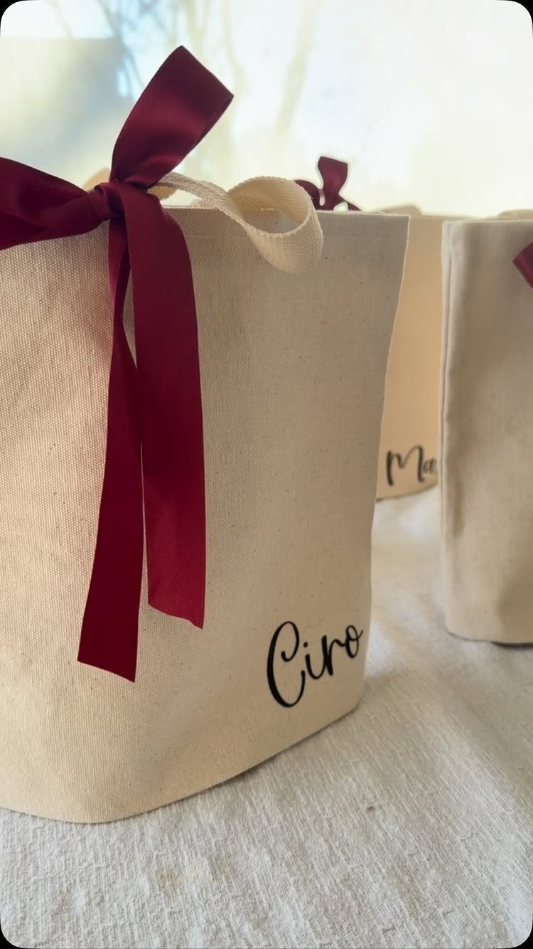 Cream canvas tote bags with personalized names and red satin ribbon bows.