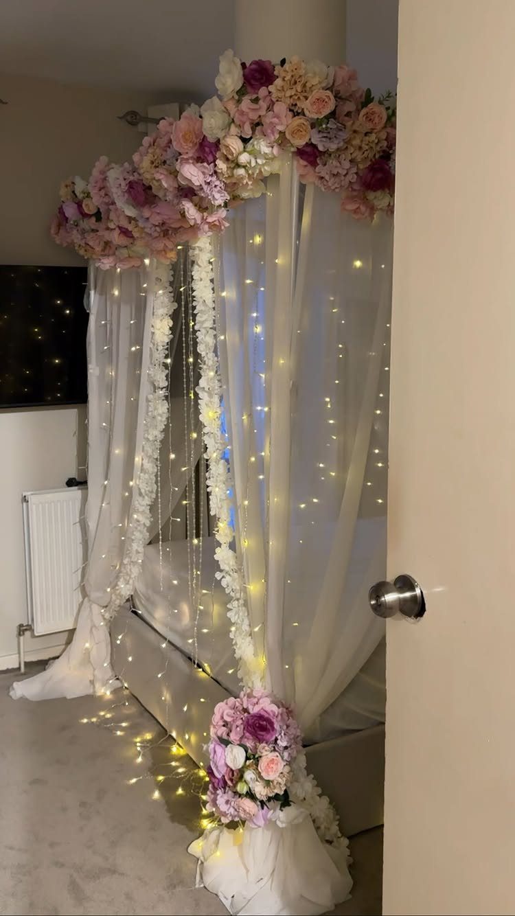 Bed with white sheer canopy, fairy lights, and pink floral arrangements for wedding night.