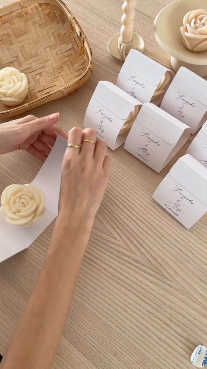 Rose-shaped candles being placed into personalized white gift boxes on a wooden table