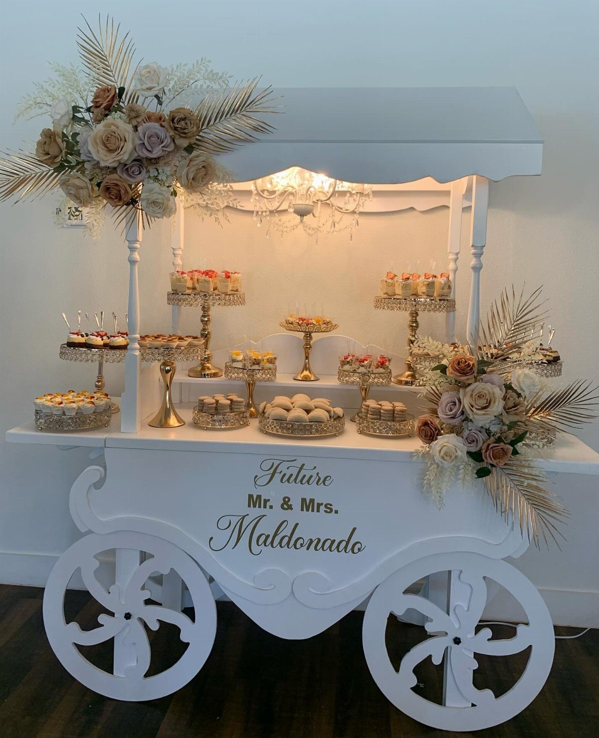 White carriage-shaped dessert cart filled with pastries and decorated with flowers and chandelier