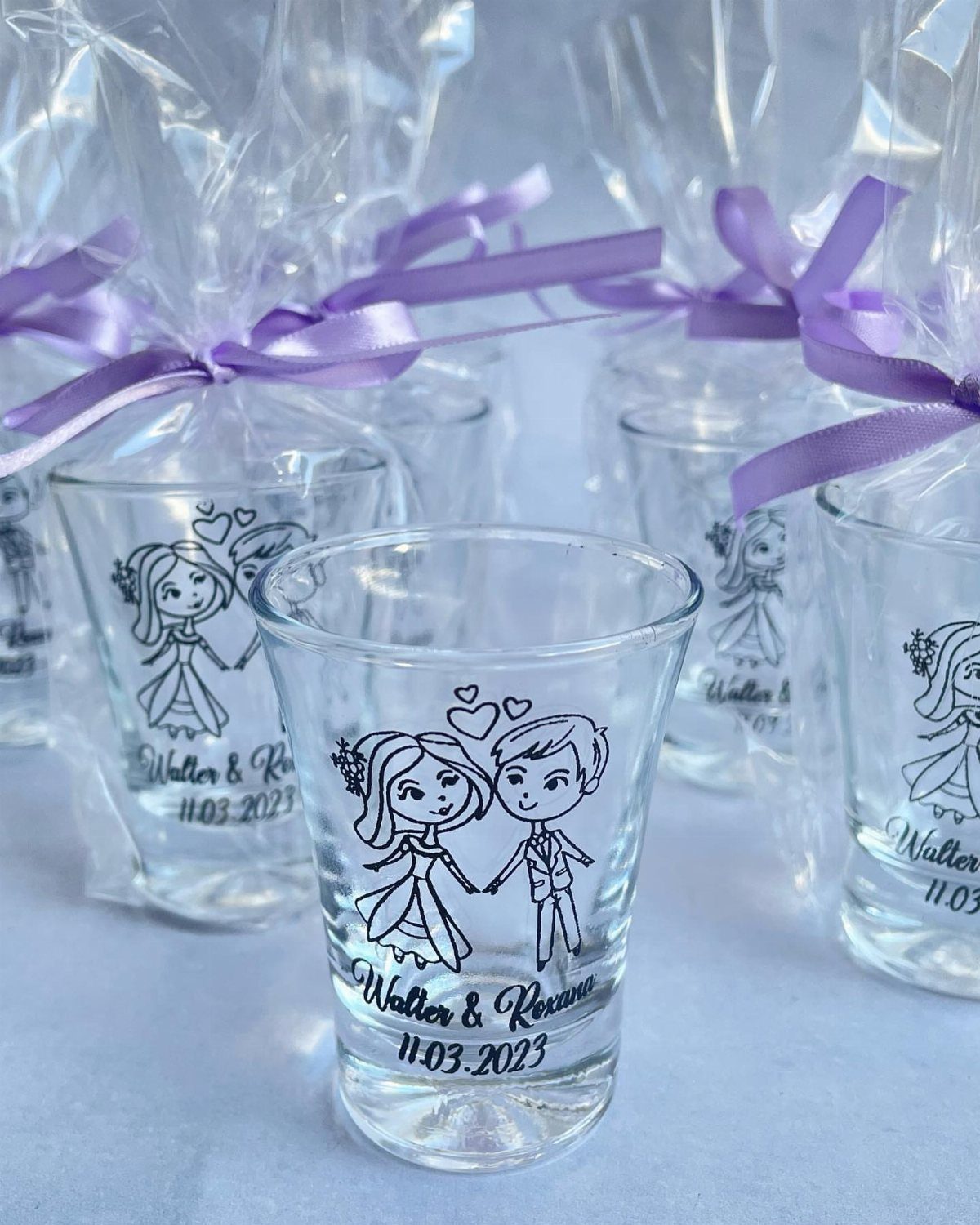 Personalized wedding shot glasses with bride and groom illustrations and purple ribbons.