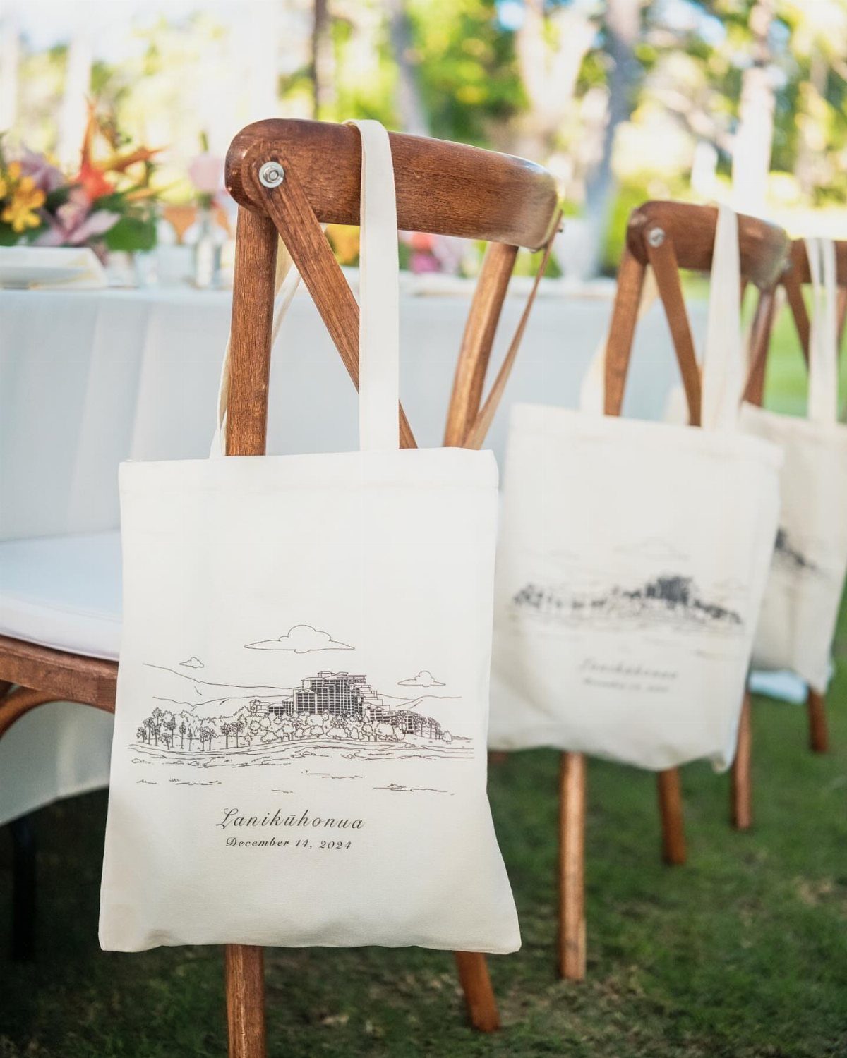 Cream canvas tote bags with custom illustrations hanging on wooden wedding chairs outdoors.