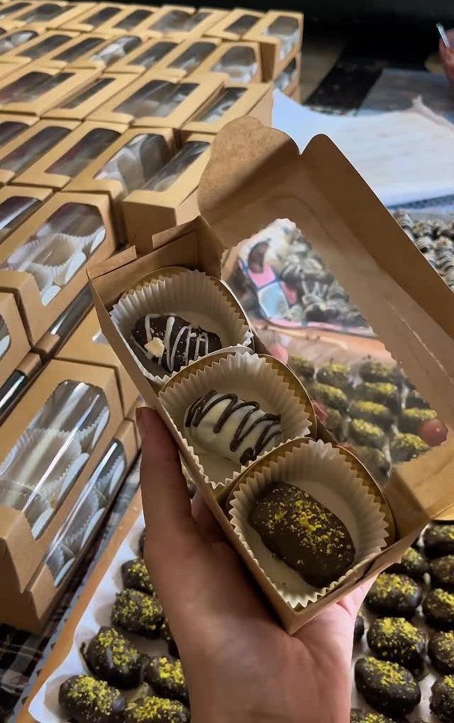 Hand holding a small brown box with three decorated gourmet chocolates inside.