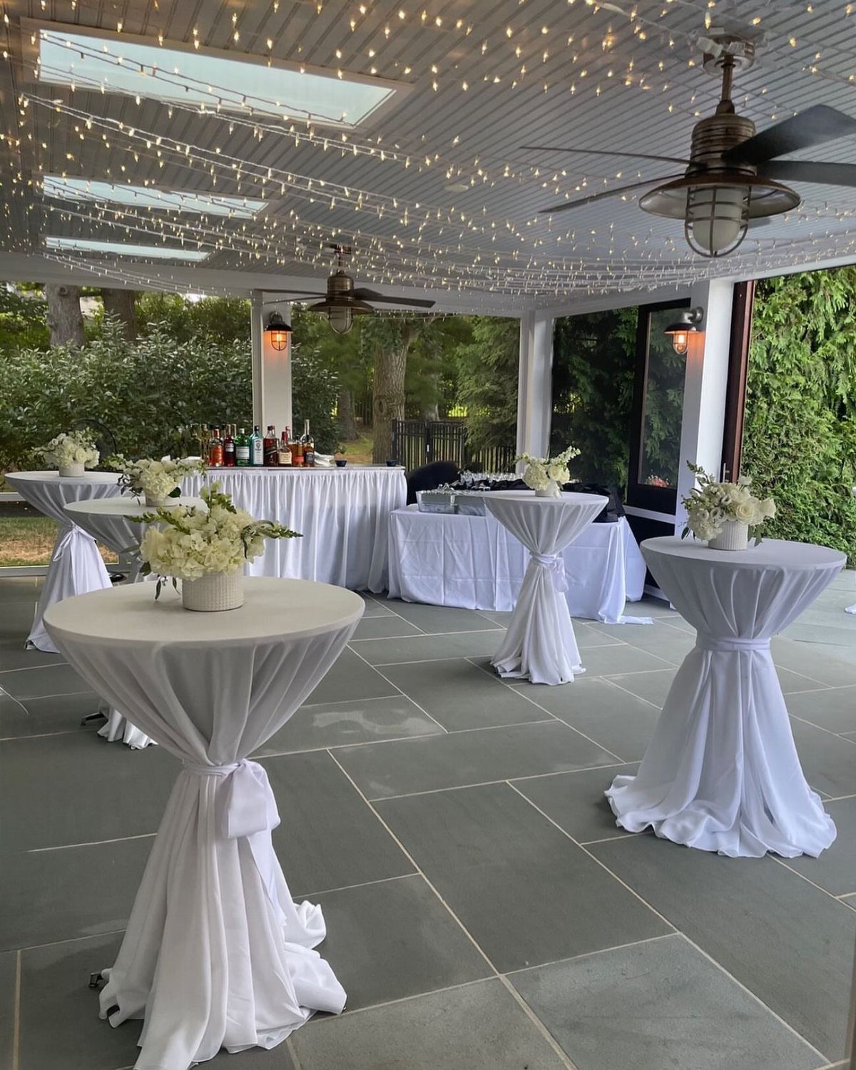 High-top cocktail tables with white linens and flowers on a lit patio.