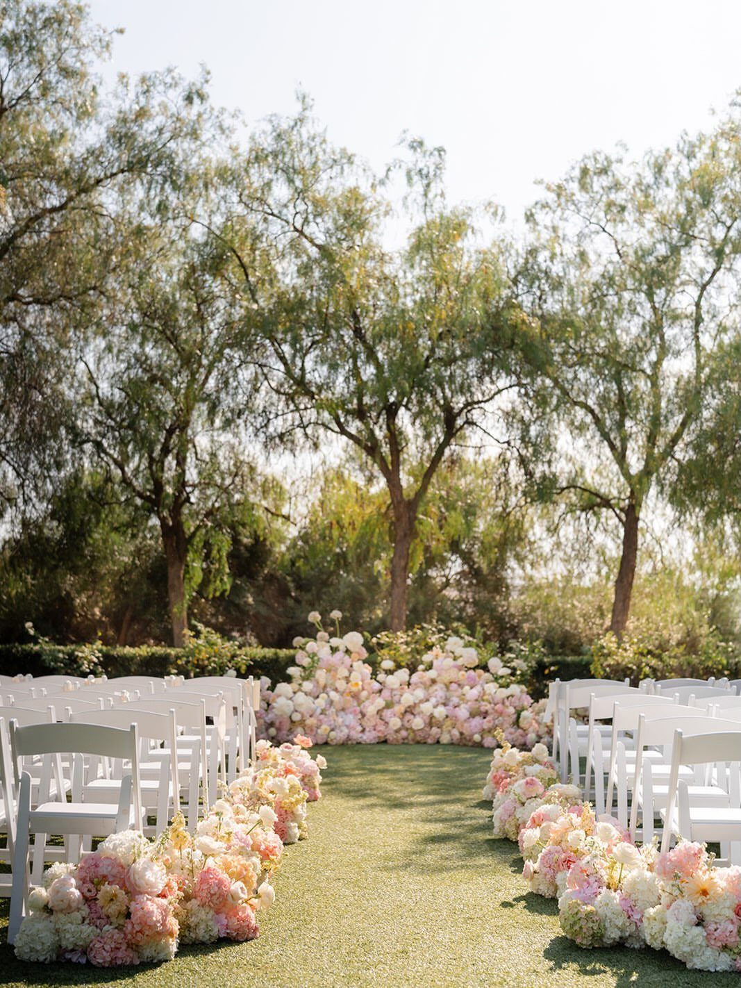 Outdoor wedding ceremony aisle with white chairs and pink and white floral arrangements.