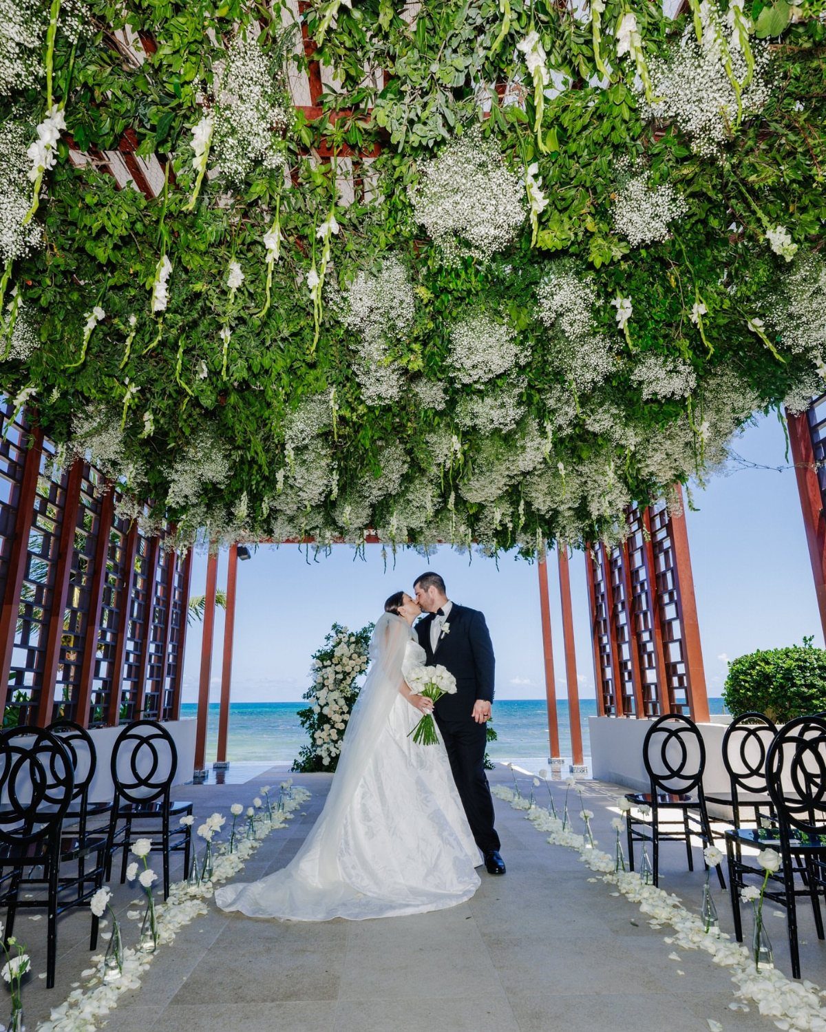 Bride and groom kissing under a dense green and white floral canopy outdoors.
