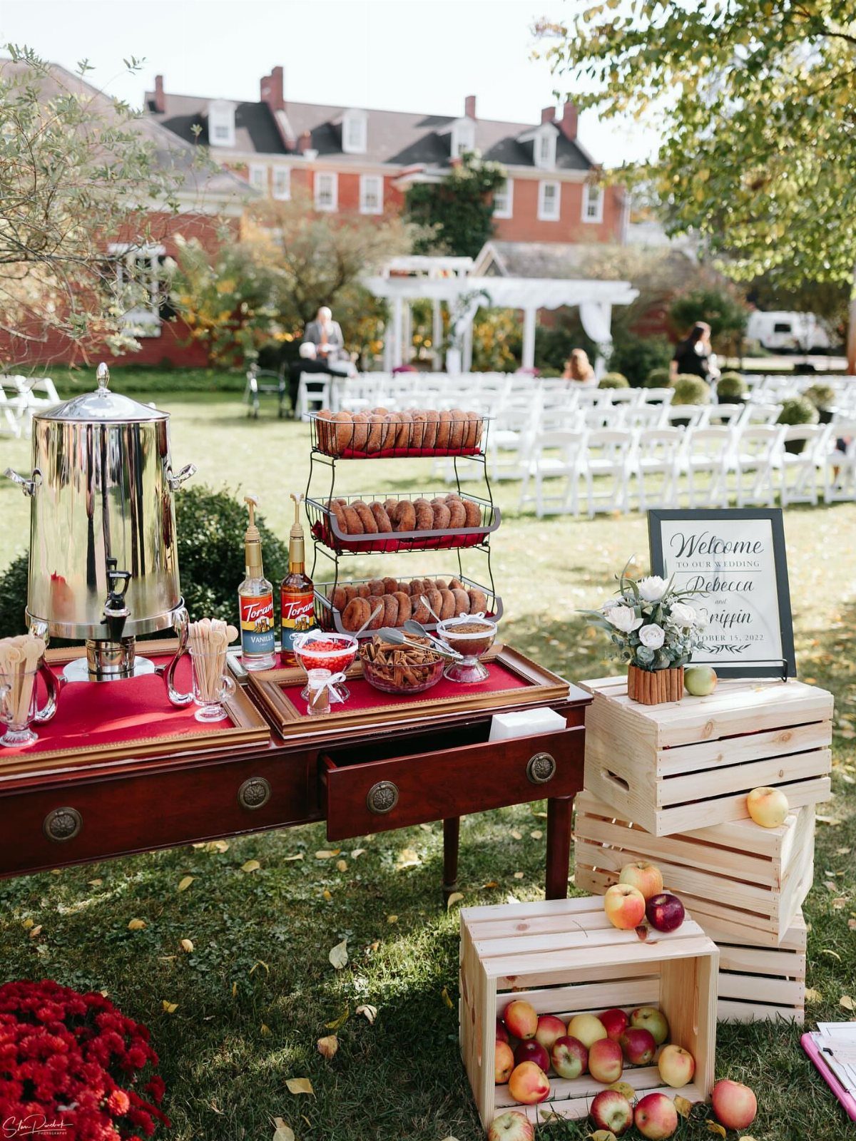 Outdoor wedding drink station with silver dispenser, donuts, and crates of red apples.