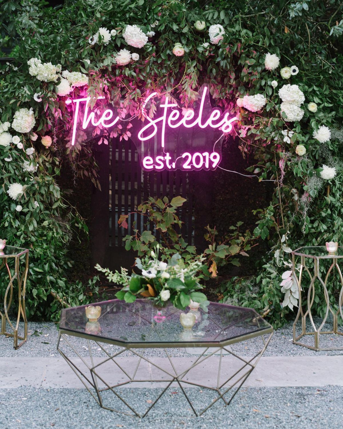 Pink neon sign with couple's name on a flower-decorated green hedge wall.