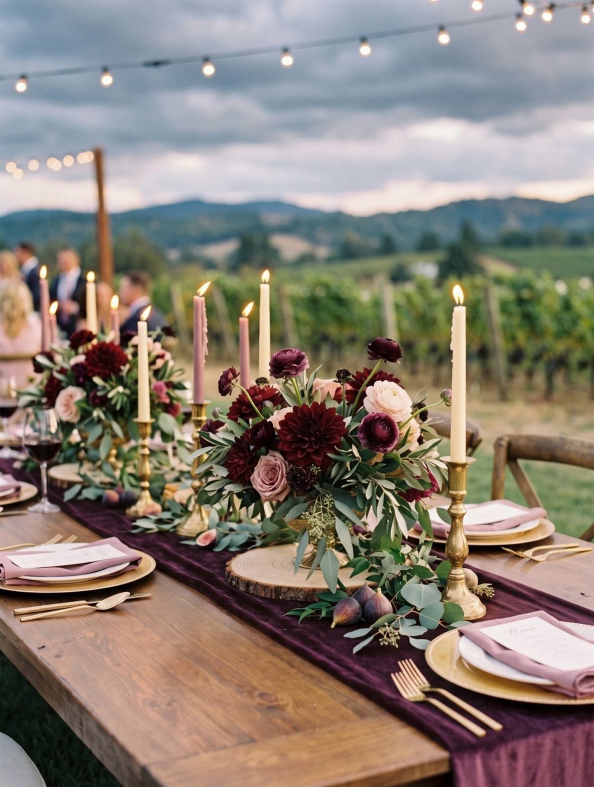 Wooden wedding table with purple runner, burgundy floral centerpieces, and gold accents outdoors.