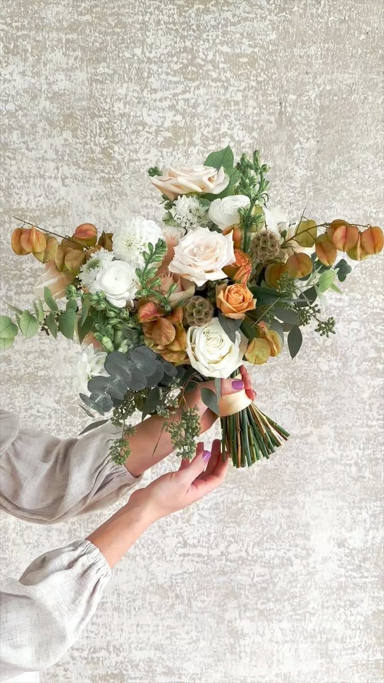 Bridal bouquet with white and peach roses and autumn foliage held by hands.