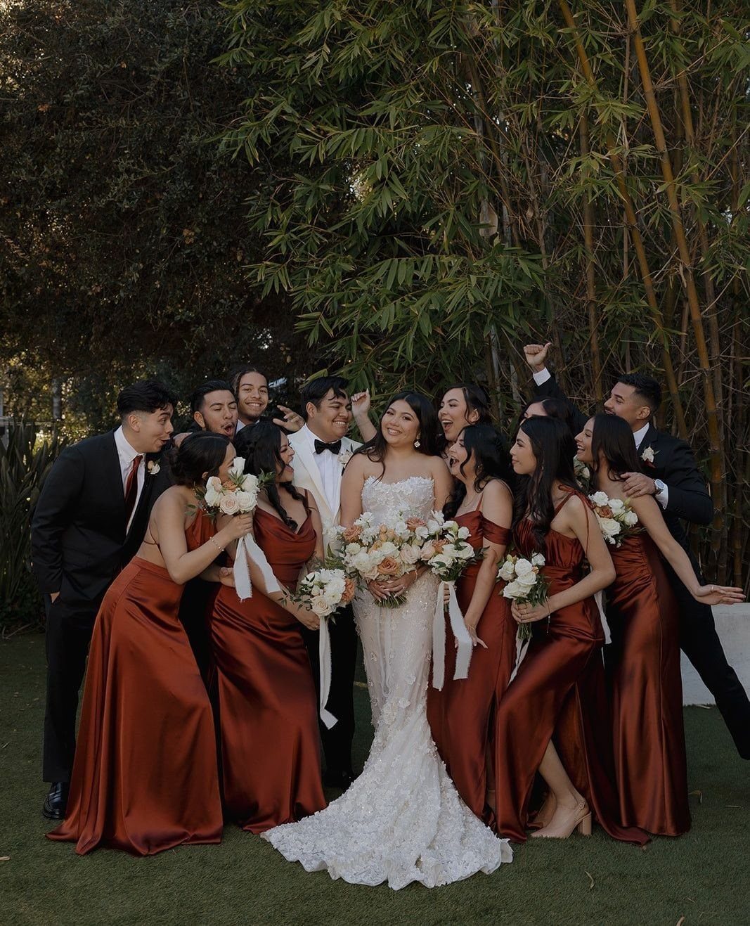 Wedding party with bridesmaids in rust satin dresses posing with the bride outdoors.